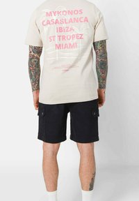 T-shirt in cotone beige con testo rosa e grafica di palma sul retro, abbinato a pantaloni cargo blu scuro con tasche laterali.