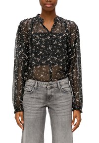 Kvinde iført en sort gennemsigtig blomstret bluse med lange ærmer og grå højtaljede jeans, der står foran en hvid baggrund.