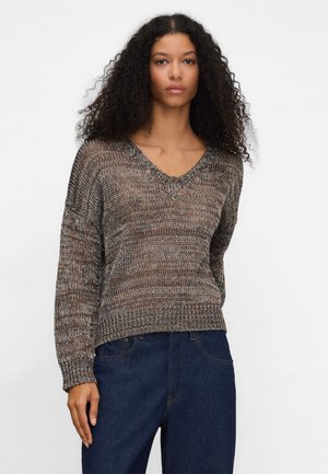 Donna con capelli ricci che indossa un maglione a V lavorato a maglia marrone e grigio e jeans blu scuro, in piedi davanti a uno sfondo semplice.