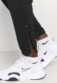 Zwarte sportbroeken met zijritsen; gecombineerd met witte mesh Nike-schoenen en zwarte logo-accenten. Witte gestructureerde sokken zijn zichtbaar.