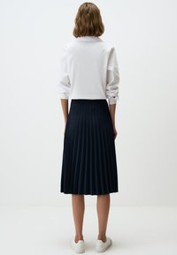 Marineblauwe geplooide rok van stof met een gladde textuur, gecombineerd met een witte blouse met kraag, gedragen met witte sneakers.
