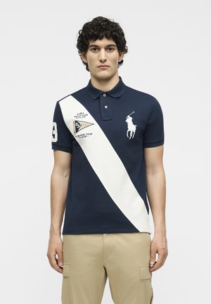 Νέος άνδρας που φοράει μπλε πόλο με λευκή διαγώνια ρίγα, μεγάλο λογότυπο παίκτη πόλο και μπεζ παντελόνι cargo, στέκεται σε απλό φόντο.