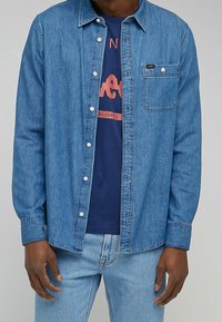 Man in een blauwe spijkerblouse over een marineblauw graphic t-shirt en lichte spijkerbroek, staand tegen een effen achtergrond.