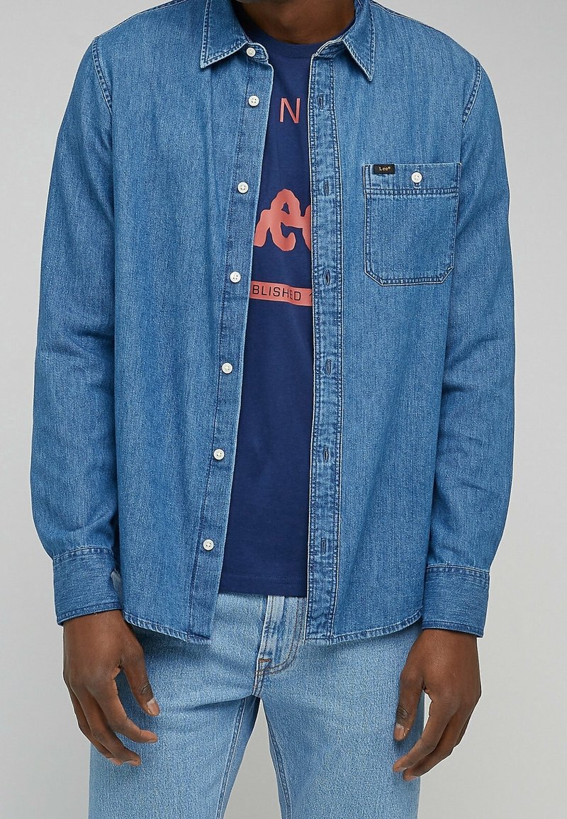 Man in een blauwe spijkerblouse over een marineblauw graphic t-shirt en lichte spijkerbroek, staand tegen een effen achtergrond.