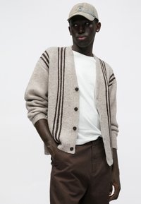 Beżowy kardigan w ciemnobrązowe paski, zapinany na guziki, z prążkowanymi mankietami, noszony na białej koszulce, zestawiony z brązowymi spodniami i beżową czapką.