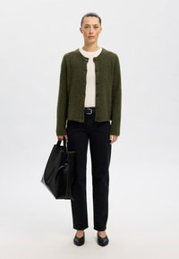 Grønn strikket cardigan med rund hals og seks knapper, båret over en hvit skjorte. Kombinert med mørke jeans og en svart handlenett.