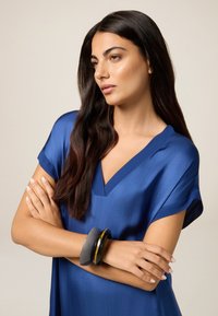 Donna con lunghi capelli scuri che indossa un vestito blu a V e bracciali spessi neri e grigi, braccia incrociate, che guarda contemplativa di lato.
