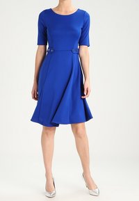 Robe bleu vif à manches courtes avec un corsage ajusté et une jupe évasée, comportant deux boutons décoratifs à la taille. Tissu au toucher doux.