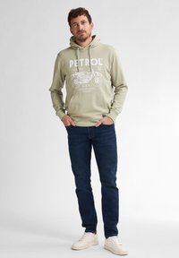Sudadera con capucha en un verde oliva claro con estampado gráfico blanco; combinada con jeans de un azul oscuro y zapatillas beige. Ajuste casual, tejido suave.