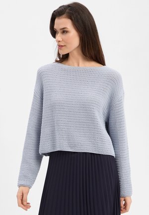 Frau trägt einen hellgrauen, strukturierten Strickpullover mit langen Ärmeln und weitem Ausschnitt, kombiniert mit einem schwarzen, plissierten Rock.