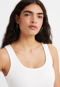 Hvid ribbet tanktop med rund udskæring og tynde stropper. Modellen har bølget mørkt hår og bærer små guldøreringe.