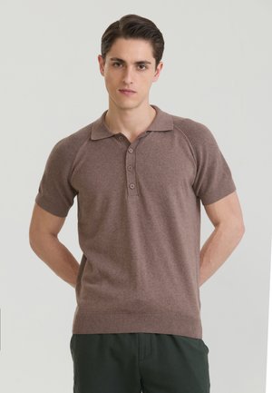 Fiatal férfi, sötét hajjal, barna rövid ujjú, gombos galléros pólót és sötét nadrágot visel, kezeit a háta mögött tartva áll.