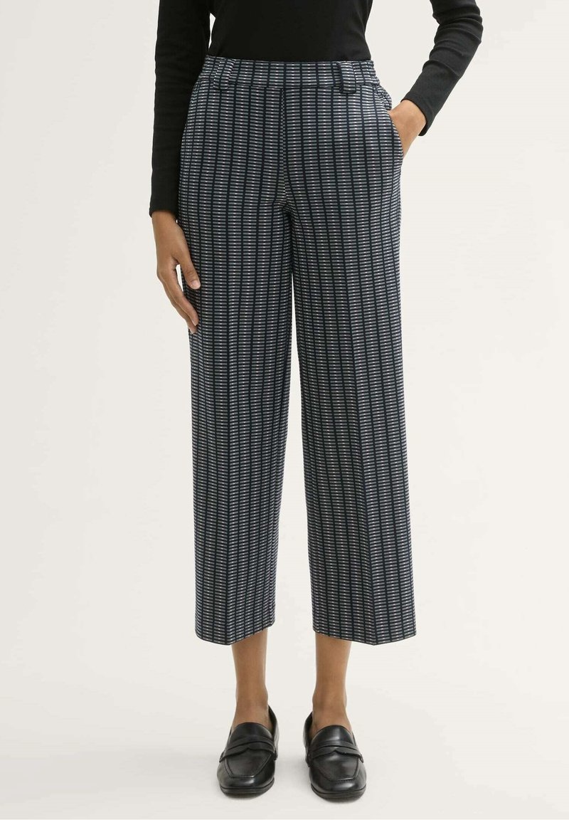 Personne portant un pantalon large court à motifs noir et blanc, un haut noir à manches longues et des mocassins noirs, debout devant un fond clair.