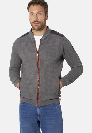 Mann mit kurzen braunen Haaren und Bart trägt eine graue, strukturierte Zip-Jacke mit orangem Besatz, blaue Jeans und steht mit den Händen in den Taschen.