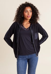 Felpa con zip di colore navy in tessuto strutturato, con accenti glitterati sulle maniche e una tasca frontale. Indossata sopra una maglietta nera con pizzo e jeans blu.