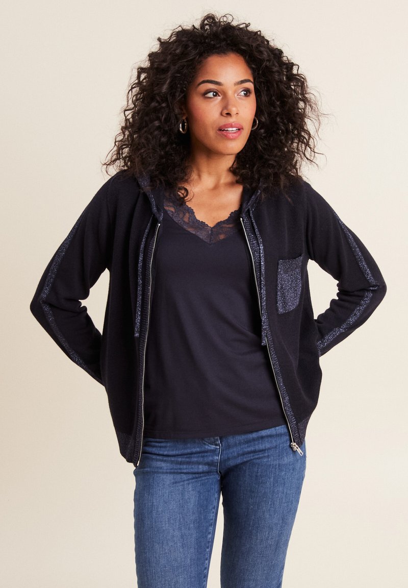 Felpa con zip di colore navy in tessuto strutturato, con accenti glitterati sulle maniche e una tasca frontale. Indossata sopra una maglietta nera con pizzo e jeans blu.