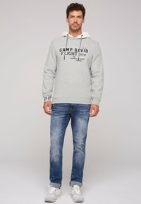 Felpa grigia chiara con un rivestimento interno bianco, con grafiche di testo scure. Abbinata a jeans blu e scarpe da ginnastica chiare, mostrando un look informale.