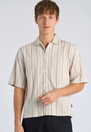 Młody mężczyzna o jasnobrązowych włosach, ubrany w kremową koszulę z krótkim rękawem, ozdobioną pionowymi czarnymi paskami, oraz czarne spodnie, stoi na tle jednolitego tła.