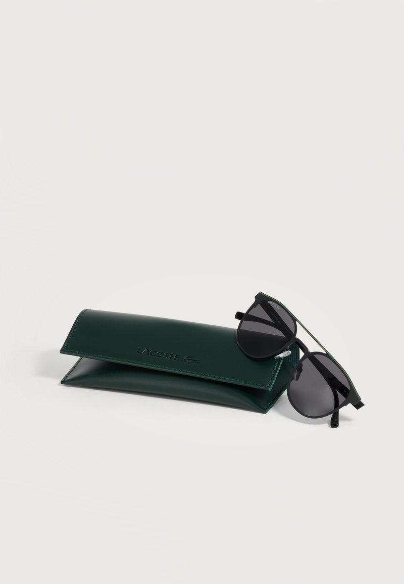 Custodia per occhiali da sole Lacoste verde scuro accanto a occhiali da sole aviator neri con lenti scure su sfondo chiaro.