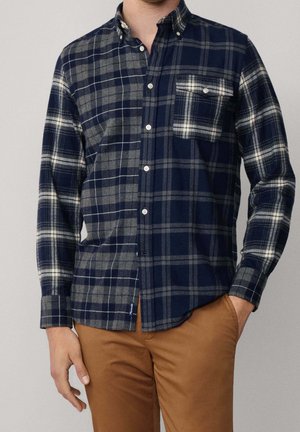 Man wearing navy and gray plaid button-up shirt with chest pocket, paired with light brown pants, standing with one hand in pocket.