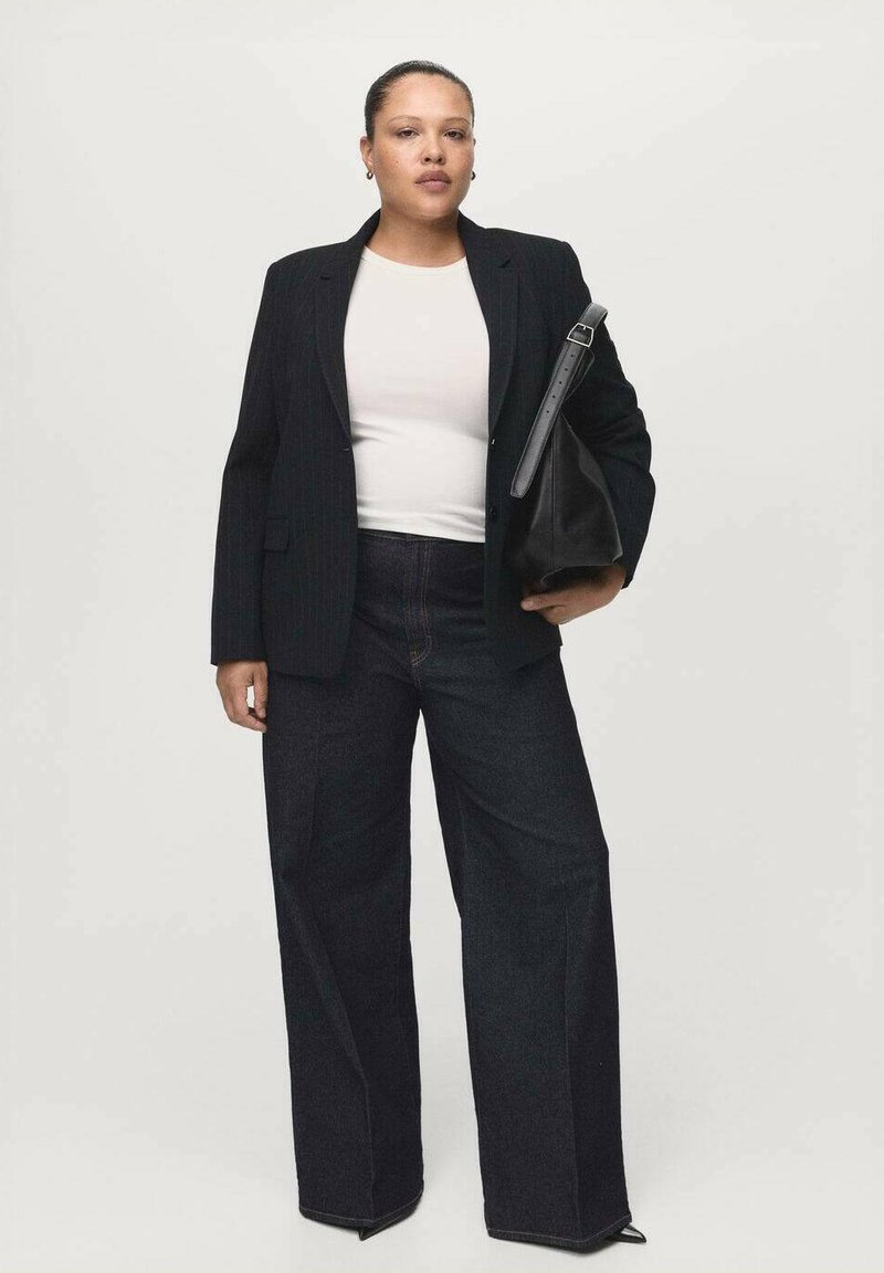 Mujer de pie con blazer negro, camisa blanca, pantalones anchos oscuros, sosteniendo un bolso grande de cuero negro sobre un fondo claro y sencillo.