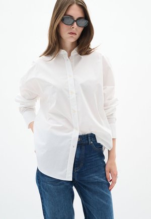 Junge Frau trägt ein übergroßes weißes Hemd mit Knöpfen, blaue Jeans und schwarze Cat-Eye-Sonnenbrille, steht vor weißem Hintergrund.