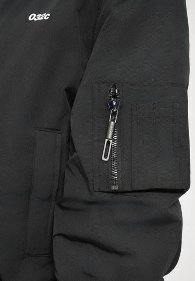 Černý rukáv bundy s kapsou na zip, která má stříbrný zip s označením "032c" a malou vyšitou nášivku.