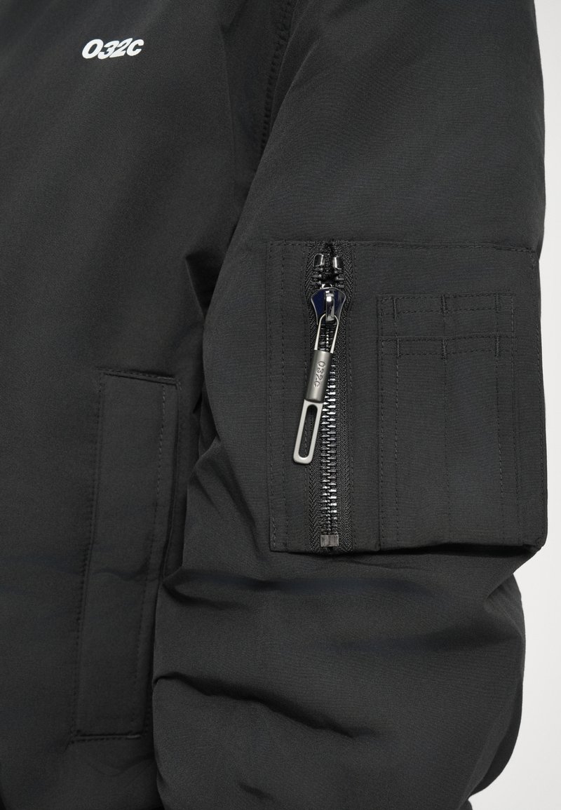 Black jacket sleeve with a zippered pocket featuring a silver zipper pull marked "032c" and a small stitched patch detail.