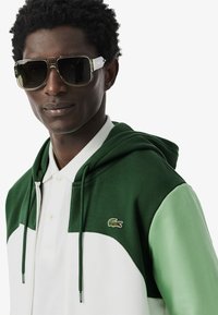 Groene en witte hooded sweatshirt met een quarter-zip ontwerp, voorzien van een Lacoste-logo. Grote donkere zonnebril met een beige accent maken de look compleet.