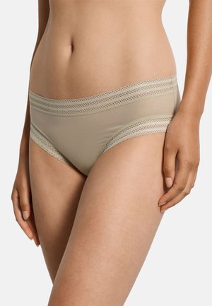 Kvinde iført beige hipster-stil undertøj med blondekant ved linningen og benåbningerne, stående mod en ensfarvet baggrund.