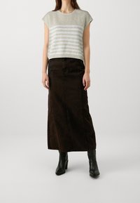 Kobieta w beżowej i białej paskowanej dzianinowej bluzce, w długiej ciemnobrązowej spódnicy i czarnych botkach na obcasie, stoi na jednolitym tle.