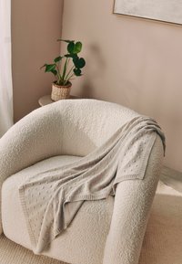 Poltrona beige e testurizzata con una forma arrotondata. Una coperta leggera grigia lavorata a maglia con punti in rilievo poggia sul bracciolo. Pianta verde in un vaso intrecciato nelle vicinanze.