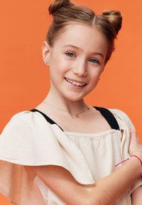 Jasbeżowa sukienka z czarnymi akcentami, falbaniastymi rękawami, delikatnym złotym naszyjnikiem i kolorowymi kolczykami przeciw pomarańczowemu tłu.