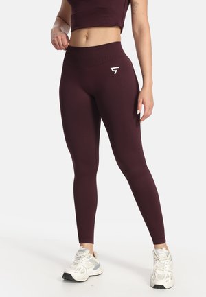 Žena nosiaca tmavofialové legíny s vysokým pásom a crop top so bielymi športovými teniskami, stojaca pred jednoduchým svetlým pozadím.