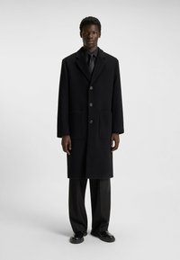Black wool coat features a notched lapel, two front pockets, and four buttons. Paired with black trousers and leather shoes.