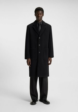 Black wool coat features a notched lapel, two front pockets, and four buttons. Paired with black trousers and leather shoes.