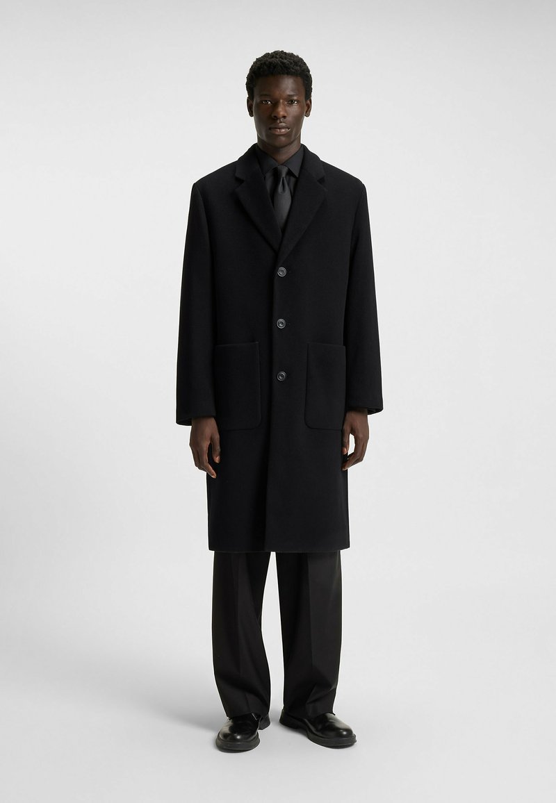 Black wool coat features a notched lapel, two front pockets, and four buttons. Paired with black trousers and leather shoes.