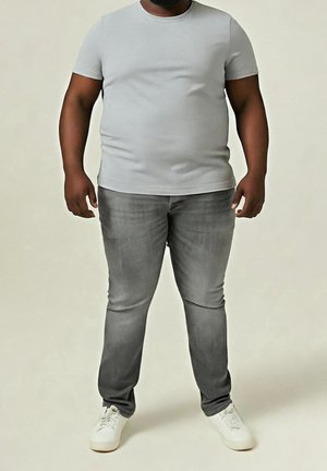 Mand iført en tætsiddende lysegrå t-shirt, falmede grå jeans og hvide sneakers stående mod en neutral baggrund.