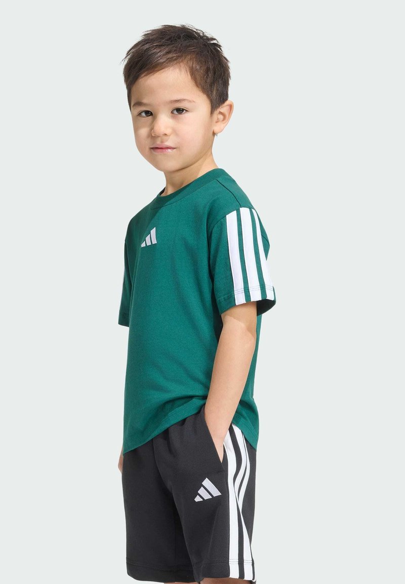 Jeune garçon debout, les mains dans les poches, portant un t-shirt vert et un short noir avec des rayures blanches sur les manches et les côtés.