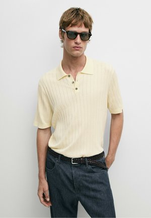 Mann trägt hellgelbes kurzärmeliges geripptes Poloshirt, dunklen gewebten Gürtel, dunkle Jeans und schwarze Sonnenbrille vor schlichtem hellem Hintergrund.