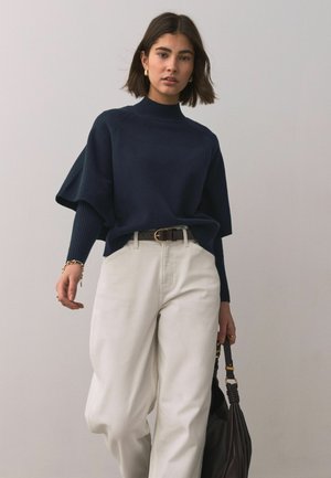 Femme portant un pull bleu marine et un pantalon blanc, tenant un sac à main noir, debout devant un fond uni gris clair.