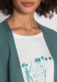 Primer plano de una persona que lleva una camisa blanca con estampado floral en verde azulado y un cárdigan texturizado en verde azulado, mostrando el cuello y los labios.