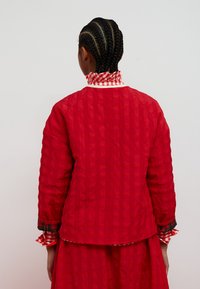 Jachetă roșie matlasată cu guler înalt cu model în carouri, cu mâneci lungi, croială lejeră și textură subtilă. Detaliu suplimentar în carouri pe manșete.