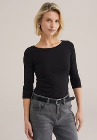Fekete, hosszú ujjú felső, kerek kivágással, szürke farmerral és fekete övvel párosítva. Simaságával és testhezálló kialakításával kiemeli a sziluettet.