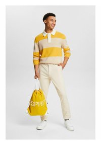 Maglione a righe gialle e crema con colletto bianco, abbinato a pantaloni beige. Zaino giallo con accenti di logo bianchi. Sneakers bianche.