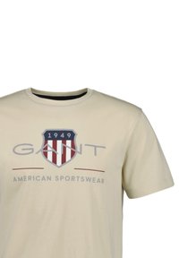 Beige t-shirt met korte mouwen met het "GANT" logo, een rood-wit gestreepte schild-embleem, "1949" en de tekst "American Sportswear" op de voorkant.