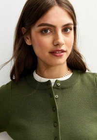 Jonge vrouw met lang bruin haar, draagt een groene cardigan over een wit shirt en glimlacht zachtjes tegen een lichte achtergrond.