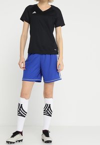 Camiseta deportiva negra de manga corta, pantalones cortos azules con rayas blancas, calcetas blancas hasta la rodilla con patrón negro y tacos con detalles en negro.