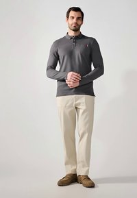 Camisa polo gris de manga larga con un pequeño bolsillo en el pecho, combinada con pantalones crema y zapatos marrones sin cordones. Diseño sencillo y corte relajado.