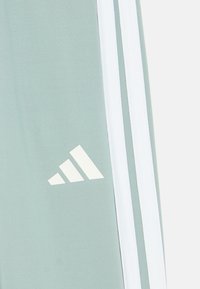 Světlé zelené sportovní kalhoty s bílými pruhy po stranách, na spodní levé noze je bílé logo Adidas. Hladká textura, zužující se střih.